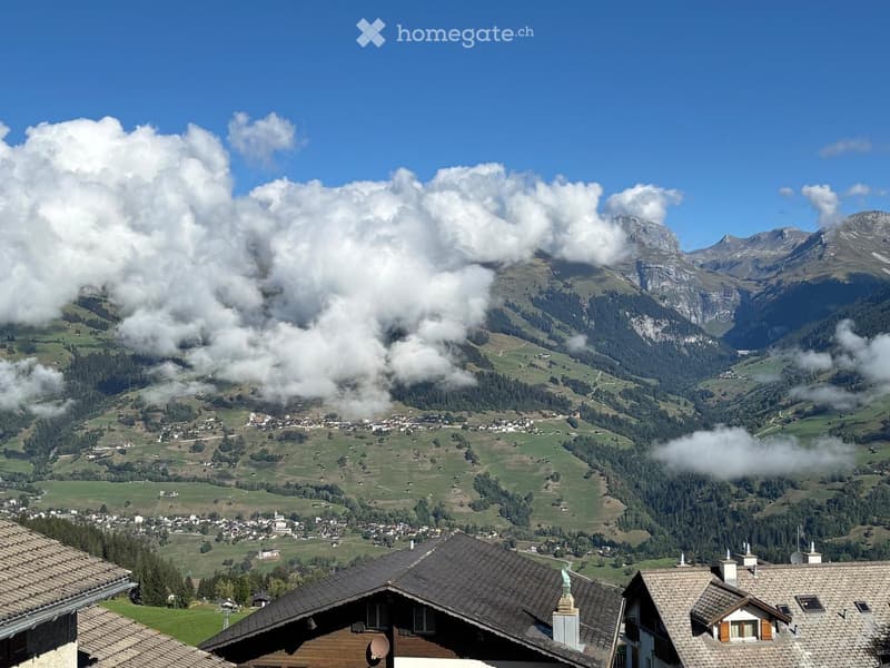 A scenic view of a village nestled in a lush green valley surrounded by towering mountains under a clear blue sky.