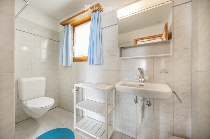Modern bathroom with white tiles, a toilet, a sink with a mirror above, a small shelf, and a window with light blue curtains.