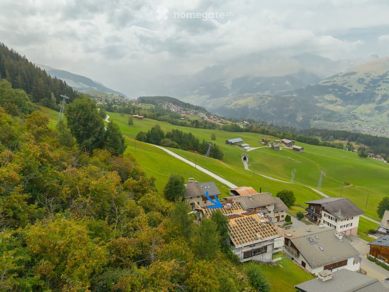 Aerial view of a rural village nestled in a green valley surrounded by mountains. Multiple houses with pitched roofs are visible, along with roads, trees, and distant mountain ranges.