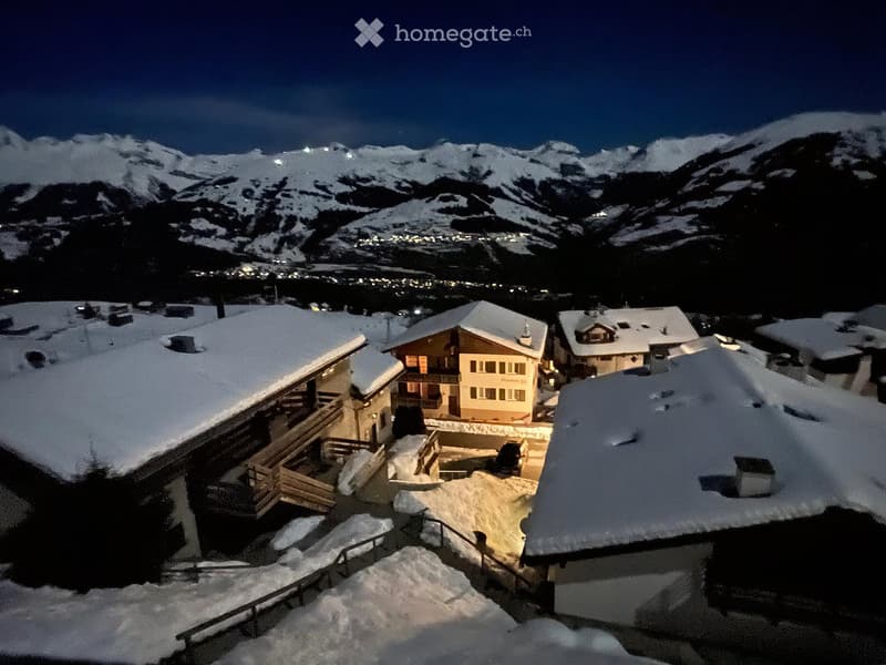 Nighttime view of a snow-covered village with traditional wooden houses and illuminated windows, surrounded by snow-capped mountains.