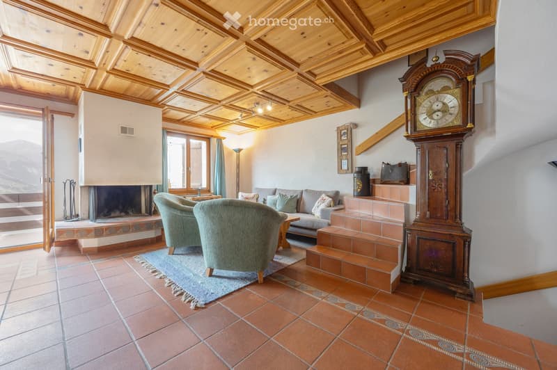 Living room with exposed wooden ceiling beams, large windows offering a mountain view, a fireplace, a sofa, armchairs, a coffee table, and a grandfather clock.