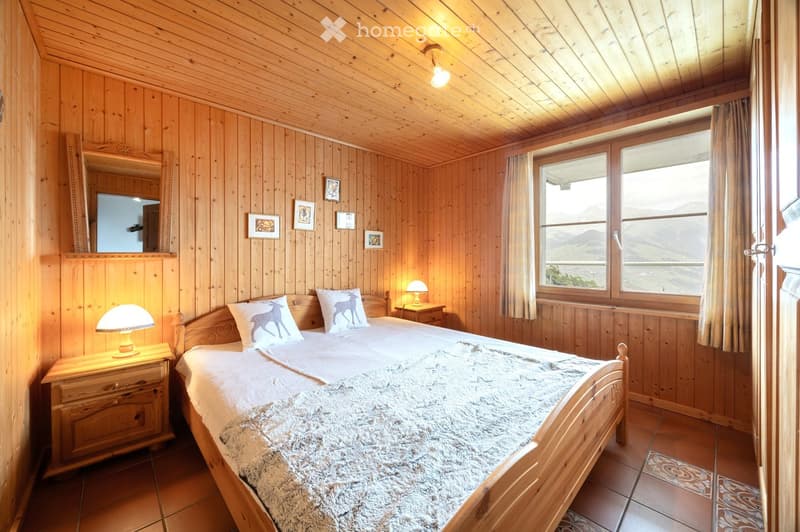 Cozy wooden bedroom with a large bed, nightstands, lamps, and a scenic mountain view through large windows.