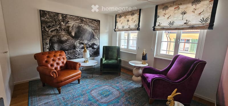 A stylish living room with a large black and white artwork, leather armchair, velvet chair, small round tables, and decorative lamps.