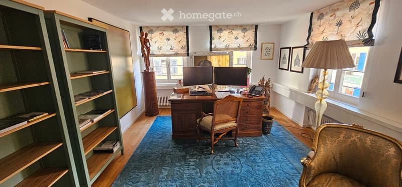 A well-furnished study or office space with a wooden desk, two computer screens, a wicker chair, a bookshelf, a table lamp, and a patterned rug.