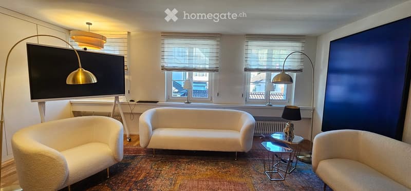 Modern living room with white walls, large windows, and a view. Features a white sofa, two armchairs, a glass coffee table, and a large television mounted on the wall. The room is illuminated by floor lamps and a ceiling light.