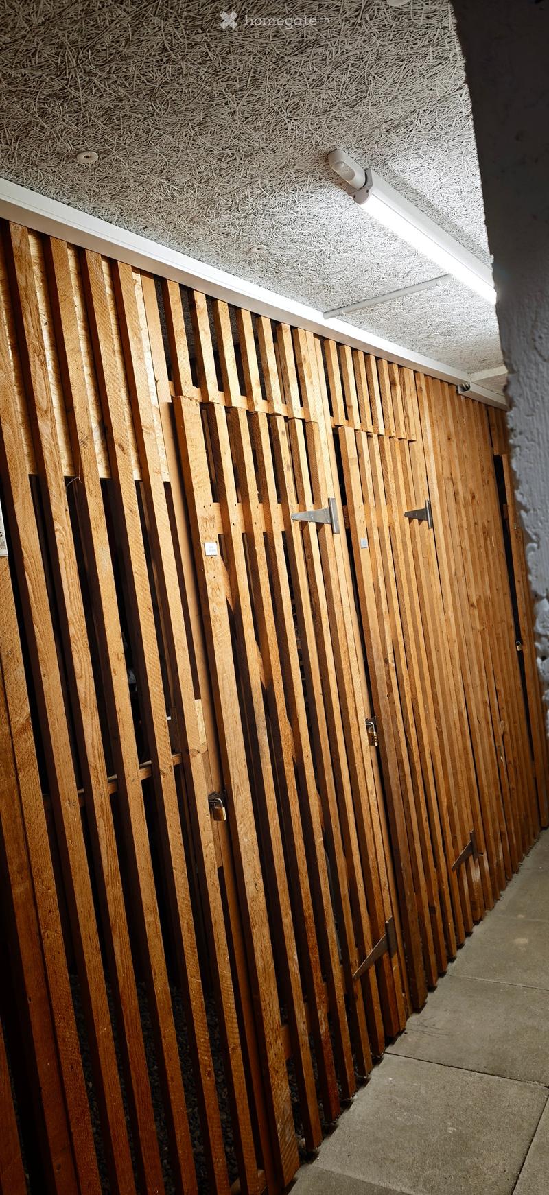 A narrow hallway with wooden slatted walls and a textured ceiling, featuring a series of metal latches and a security camera mounted on the ceiling.