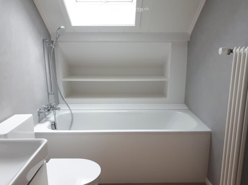 Modern, minimalist bathroom with a white bathtub, toilet, sink, and built-in shelves. Features a sleek showerhead and a radiator on the wall.
