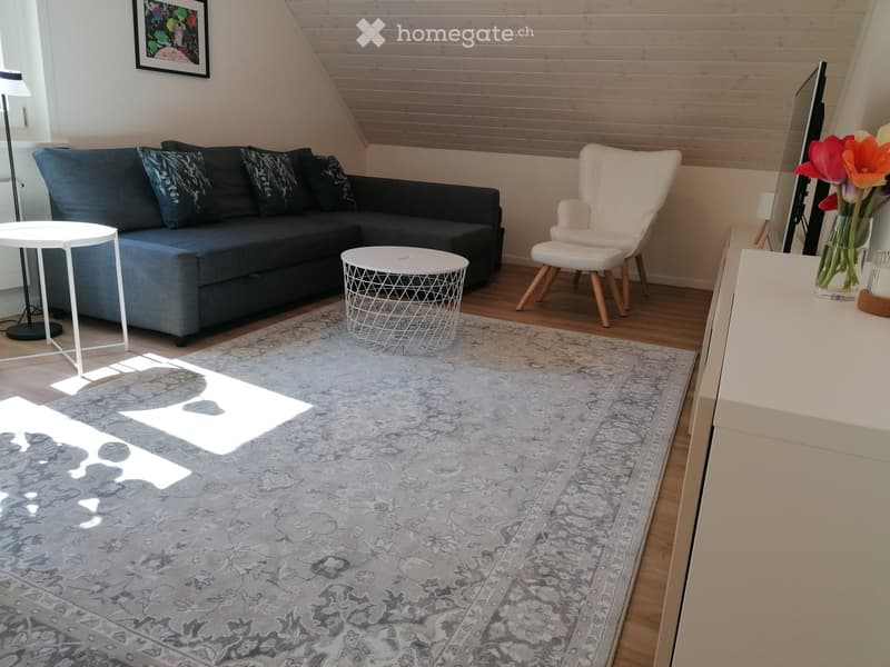 Modern living room with a grey sofa, white armchair, small round tables, a large patterned rug, and a vase with flowers on a side table.