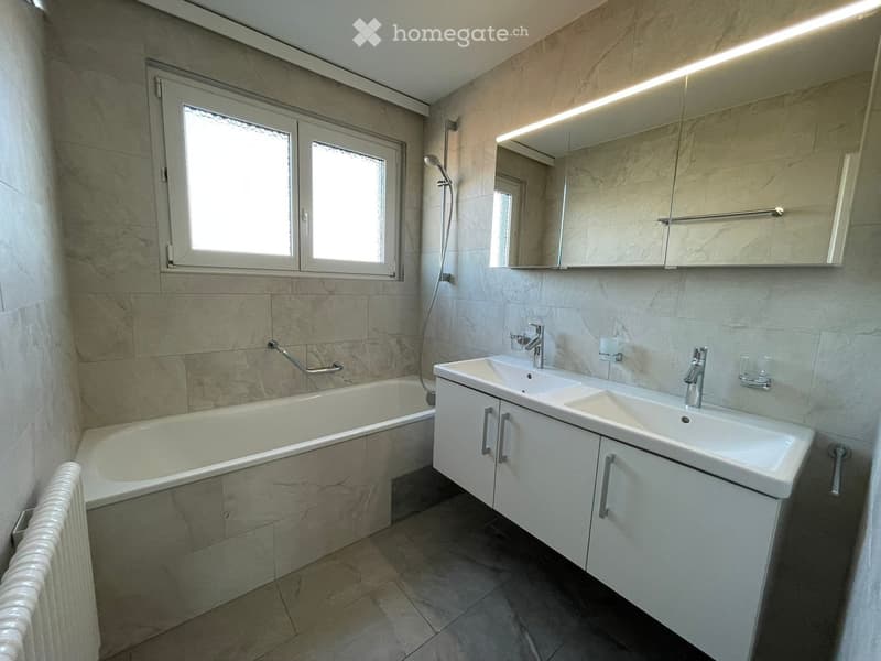 Bathroom with a bathtub, double sink, and a shower head. The walls and floor are tiled with marble effect tiles. There is a radiator on the left side.