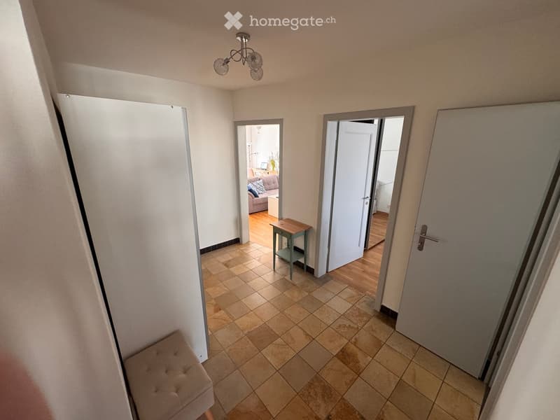 Hallway with tiled floor, white walls, and several doors leading to different rooms. A small wooden table and a bench are visible.