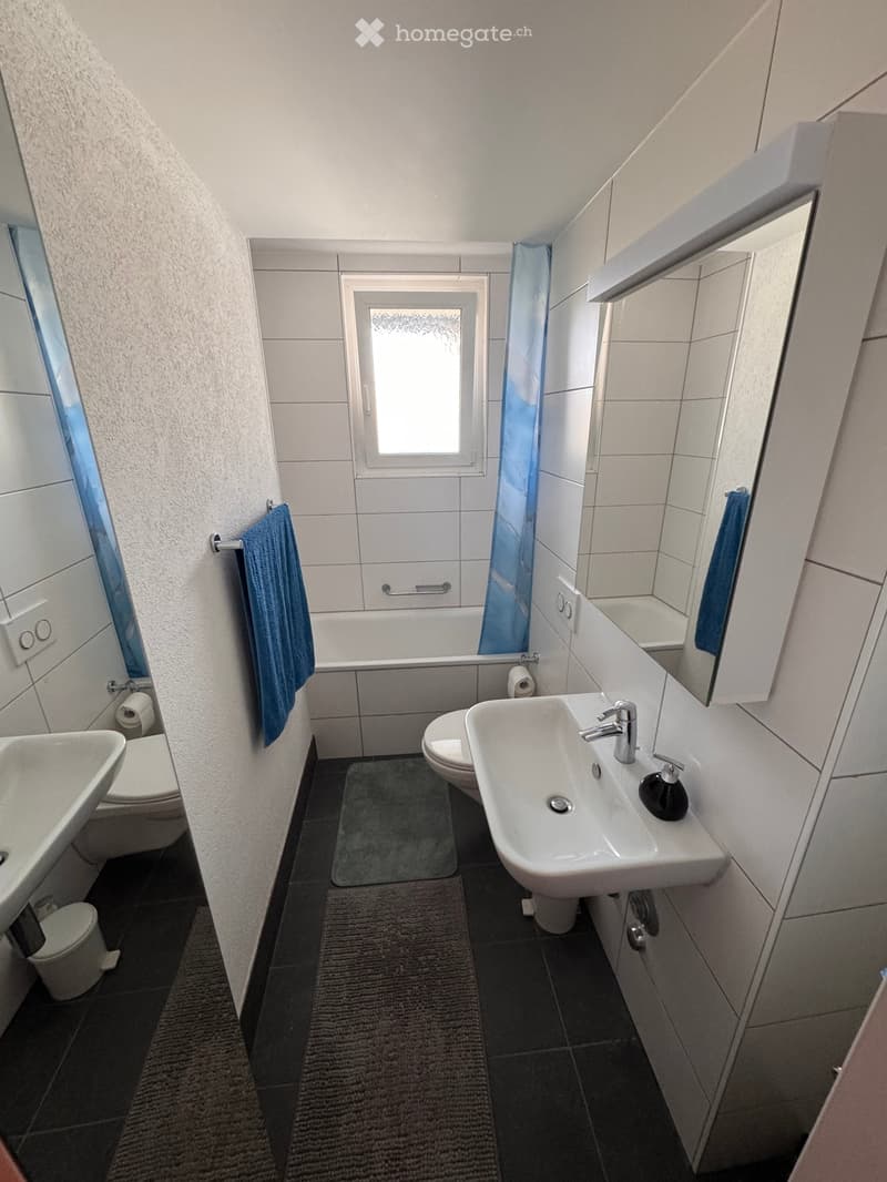 Modern bathroom with white tiles, a sink, toilet, bathtub, and a mirror. Blue towels are placed on the sides, and a small rug is on the floor.