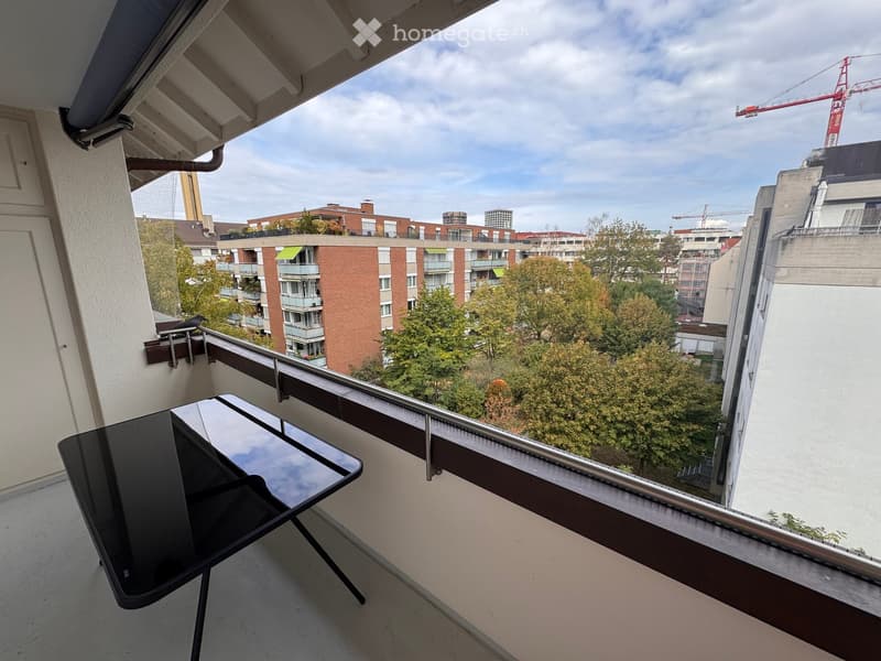 Balcony with a modern black table and chair, overlooking a cityscape with buildings and trees.