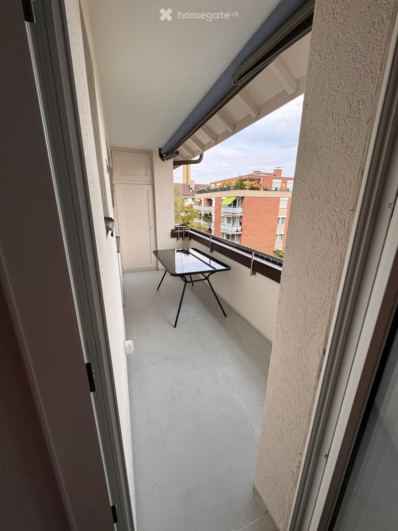 Balcony with a small black table, overlooking a residential area with buildings and greenery.