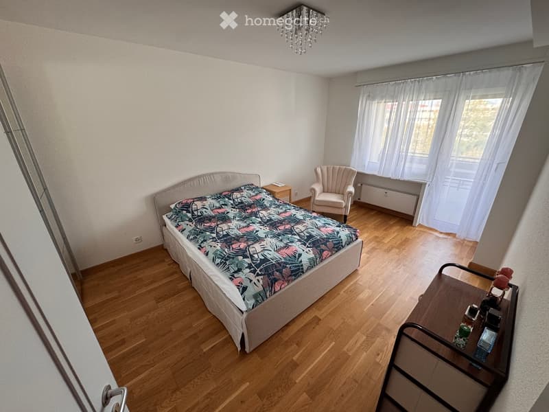 Bedroom with wooden flooring, a bed with a colorful duvet, a nightstand, a chair, and a small table with items on it. There is a window with white curtains and a chandelier on the ceiling.
