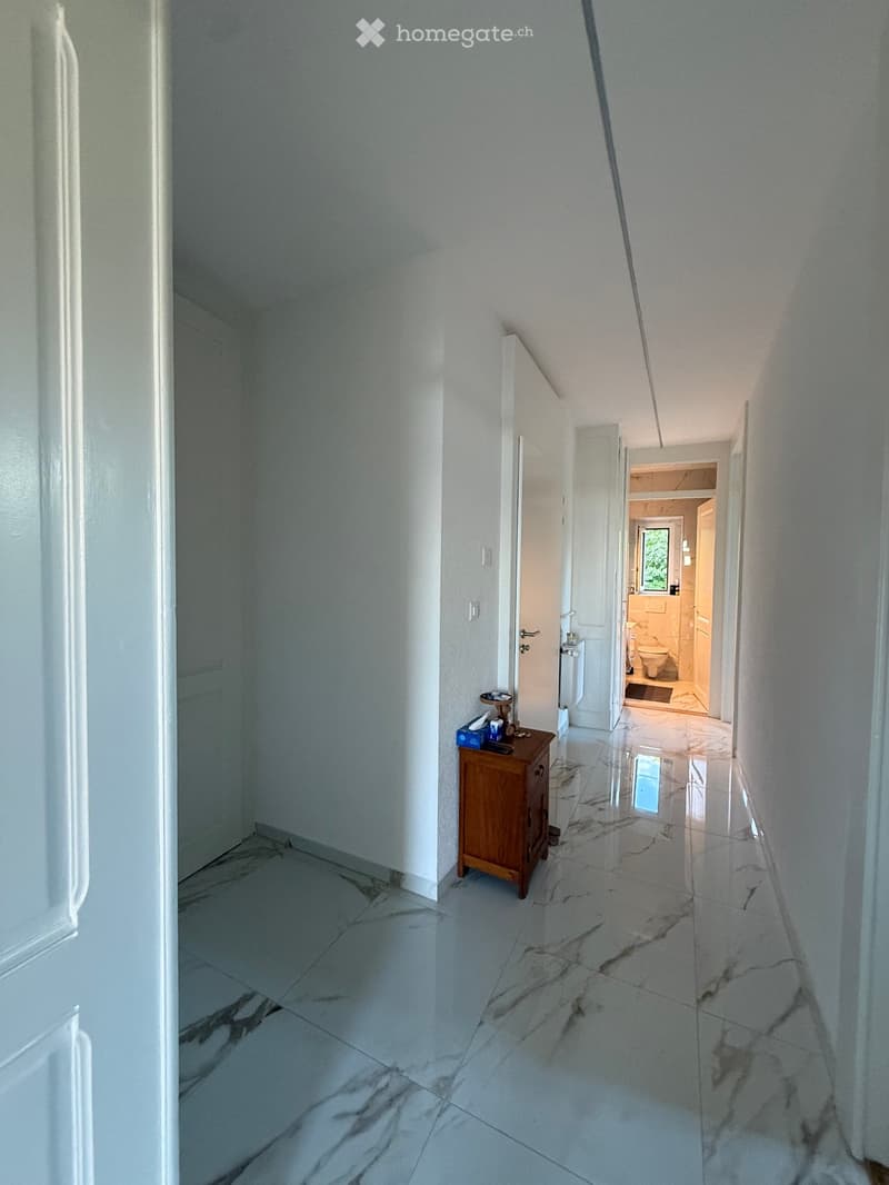 A clean, well-lit hallway with white walls and marble flooring, featuring a small wooden cabinet with items on top and an open door leading to a bathroom.