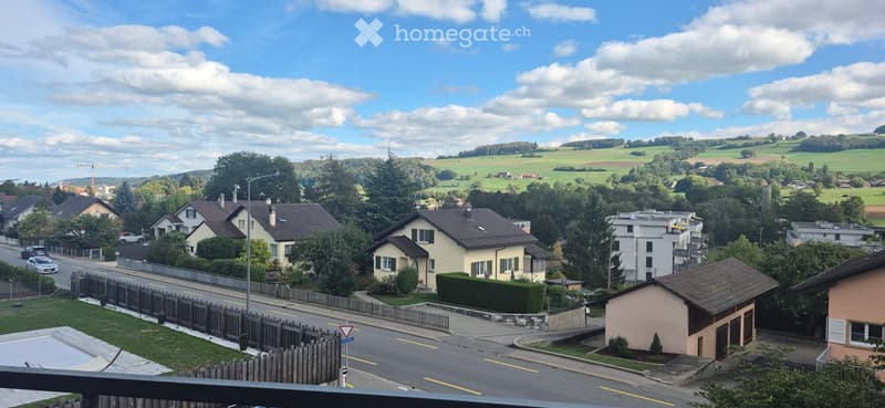 A scenic view of a suburban neighborhood with houses, trees, and a road, surrounded by green fields and hills under a partly cloudy sky.