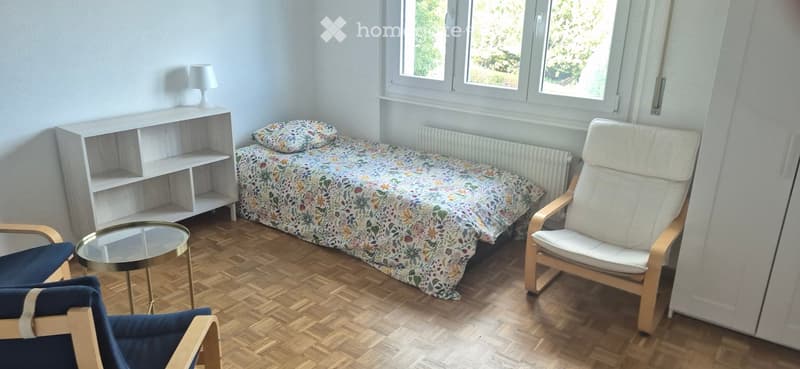 A small, well-lit bedroom with wooden flooring, a single bed with a floral duvet, a white shelf unit, a glass coffee table, and two chairs.