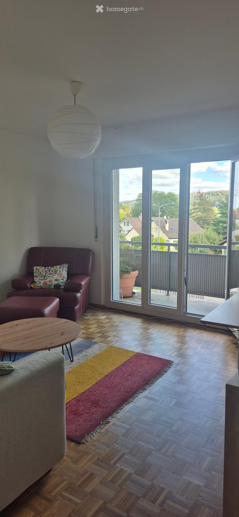 Living room with wooden flooring, a red leather sofa, a wooden coffee table, a colorful rug, and a view of the outdoors through large windows.