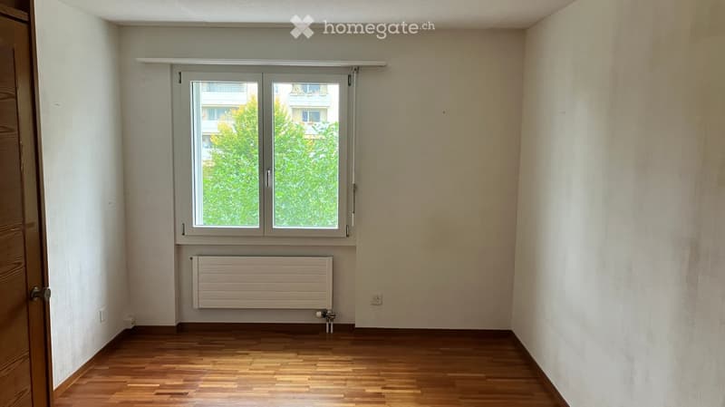 Empty room with wooden flooring, white walls, a window with a view of greenery, and a radiator below the window.