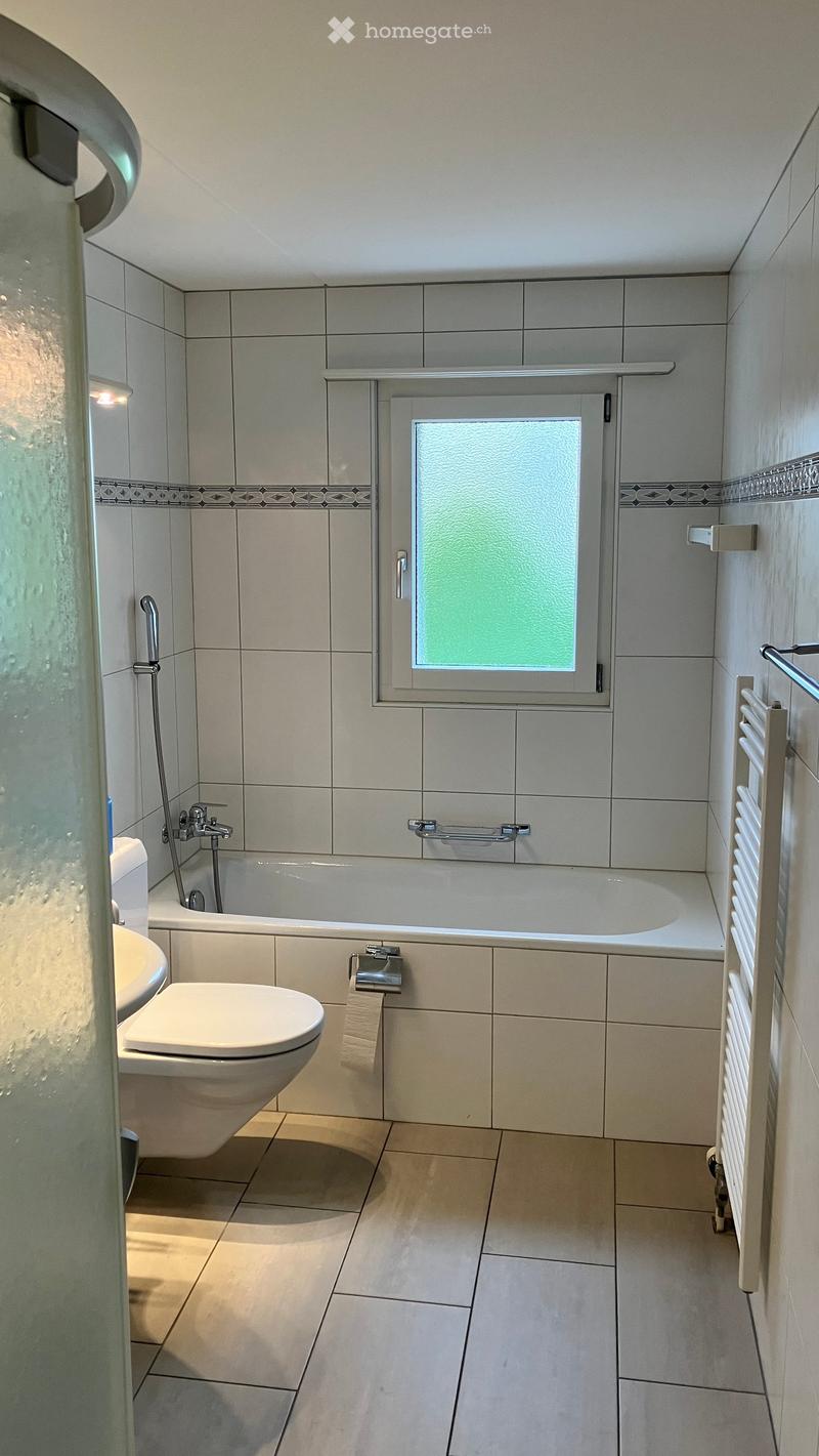 Modern bathroom with a bathtub, toilet, shower stall, radiator, and towel rack. The walls are tiled with a decorative border, and the floor is tiled in a light color.