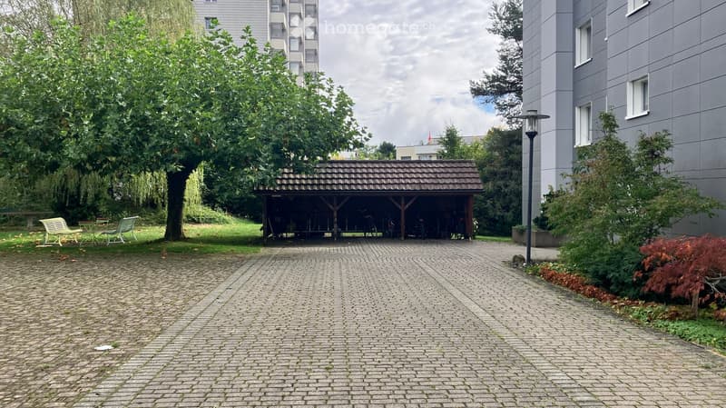 Brick-paved courtyard with a small wooden shelter housing bicycles, surrounded by trees, benches, and modern apartment buildings.