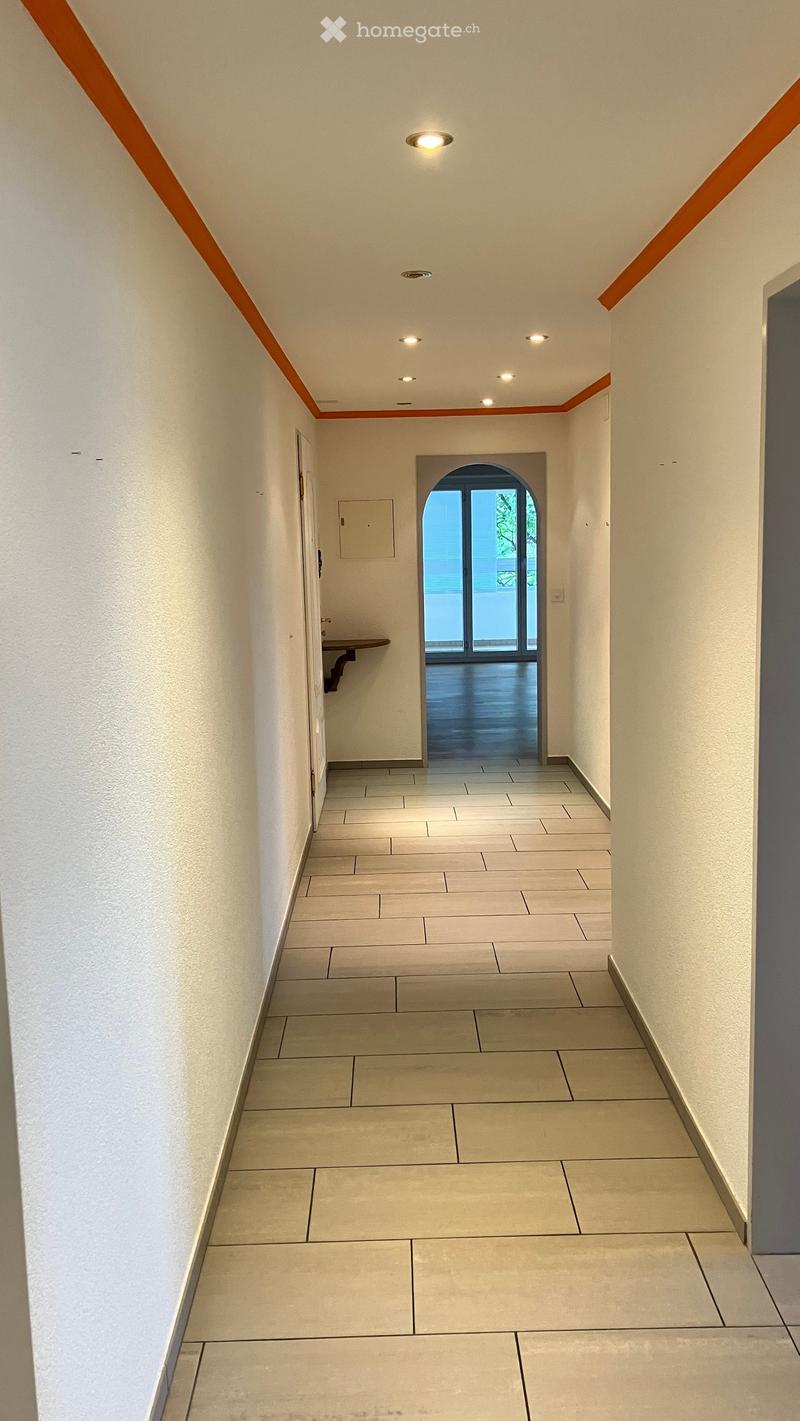 Clean, modern hallway with white walls, beige tiled floor, recessed lighting, and an arched doorway leading to a balcony area.