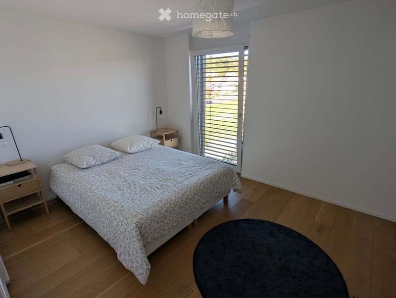 A modern bedroom with a wooden floor, a bed with a patterned duvet and pillows, a nightstand with a lamp, and a round stool near the window.