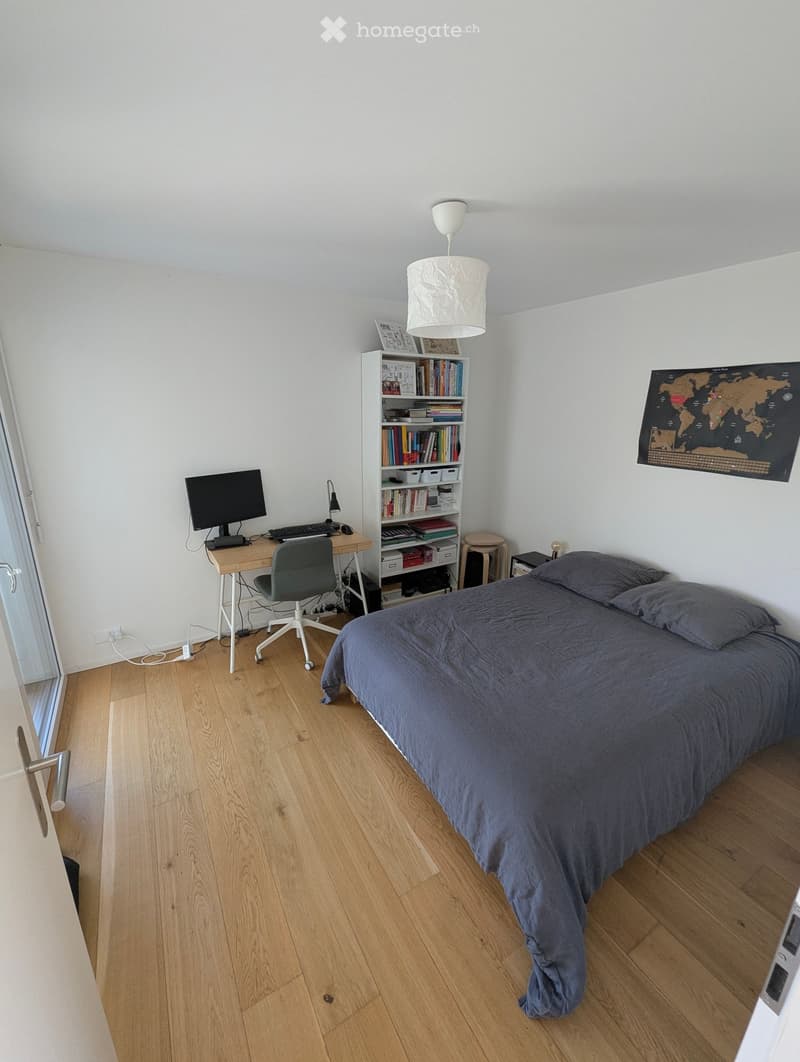 A modern bedroom with a large bed, a desk with a computer, a bookshelf, and a world map on the wall. The room features wooden flooring and white walls.