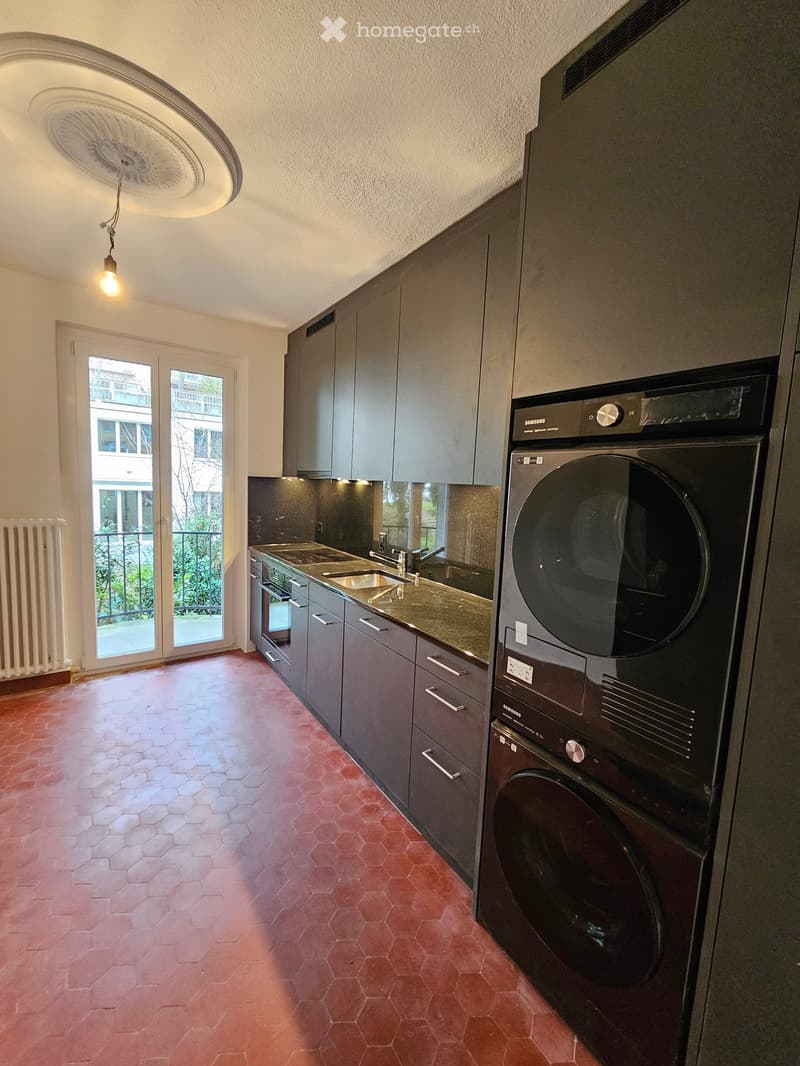 Modern kitchen with grey cabinets, a stainless steel sink, a built-in oven, and a washing machine. The floor is tiled in a hexagonal pattern, and there is a large window with a view of the balcony and greenery outside.