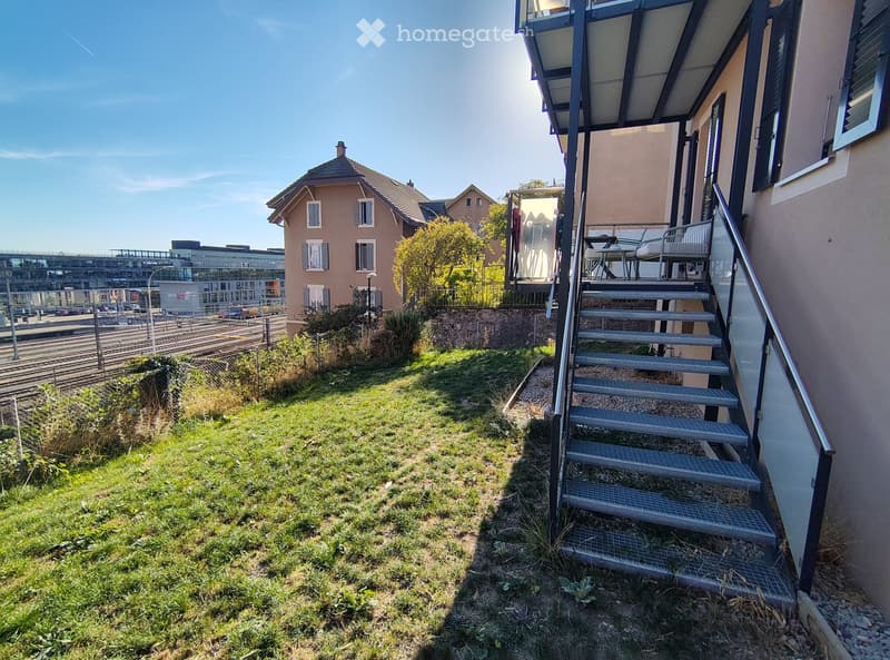 Garden area with grass, small plants, and a staircase leading to a balcony with outdoor furniture.
