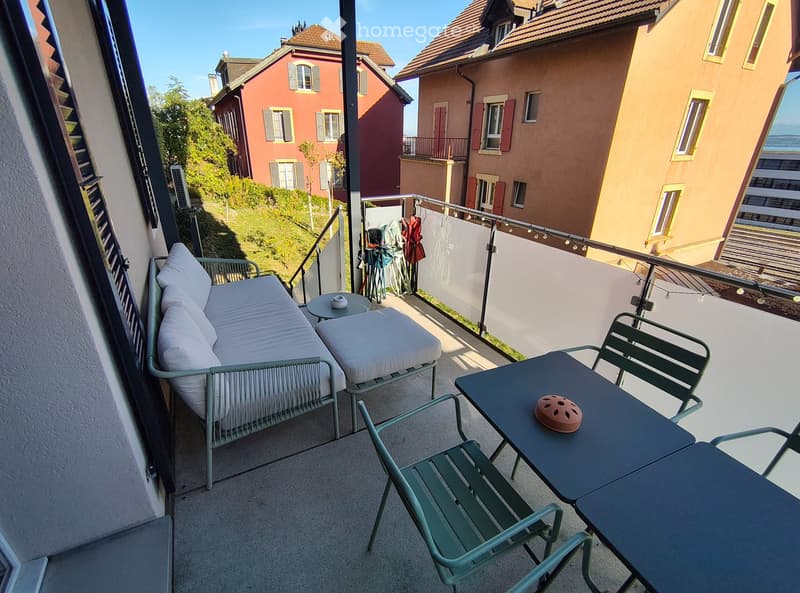Balcony with a sofa, small table, and chairs, overlooking a hillside with neighboring houses.