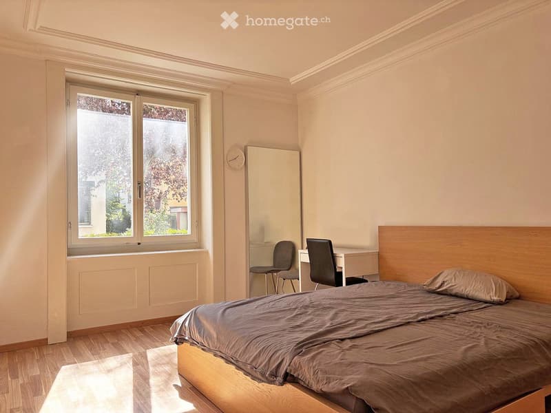 Bright bedroom with wooden flooring, large window, bed with headboard, desk with two chairs, mirror, and clock on wall.