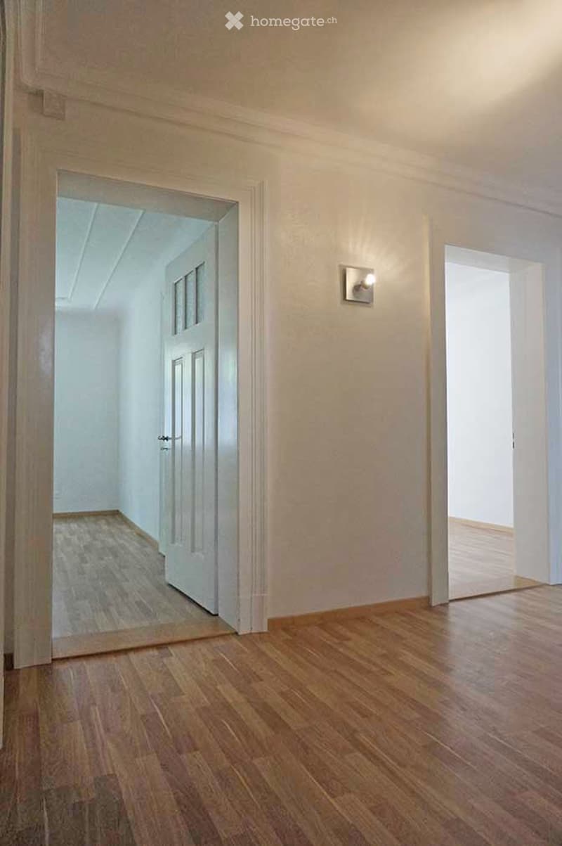 Empty hallway with wooden flooring, white walls, and a single light fixture. The hallway features an open doorway leading to another room with visible wooden flooring and white walls.