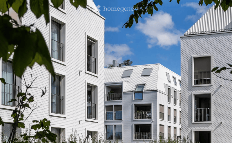 Modern white apartment building with multiple balconies and large windows, surrounded by greenery.