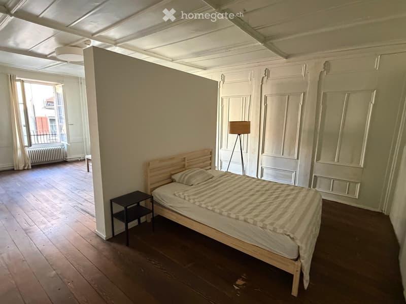 Bedroom with wooden floor, bed with striped bedding, nightstand, floor lamp, and large window with balcony access.