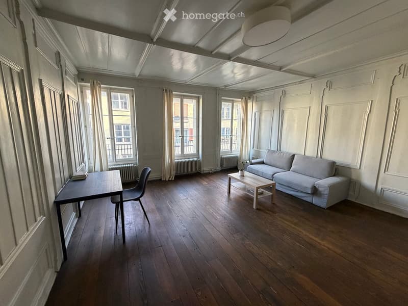 Living room with wooden flooring, large windows, a sofa, coffee table, and a small table with a chair.