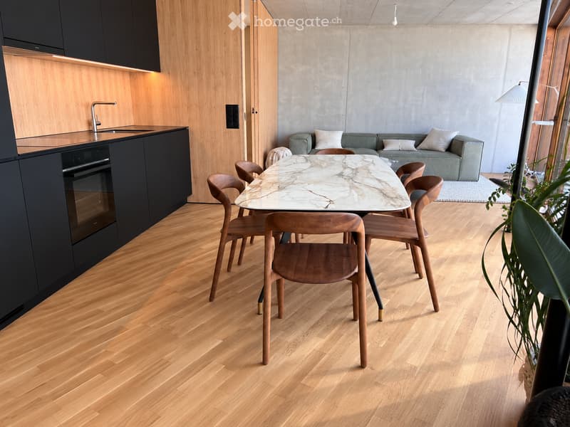 Modern open-plan living and dining area with light wood flooring, a grey sofa, a marble dining table with wooden chairs, and a sleek black kitchen with an oven and sink.