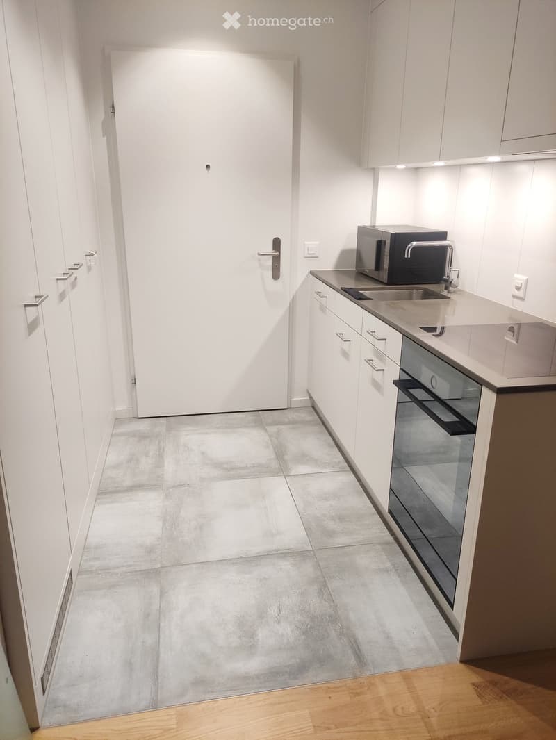 Modern kitchen with white cabinets, stainless steel sink, microwave, and oven. Concrete floor and wooden flooring transition visible.