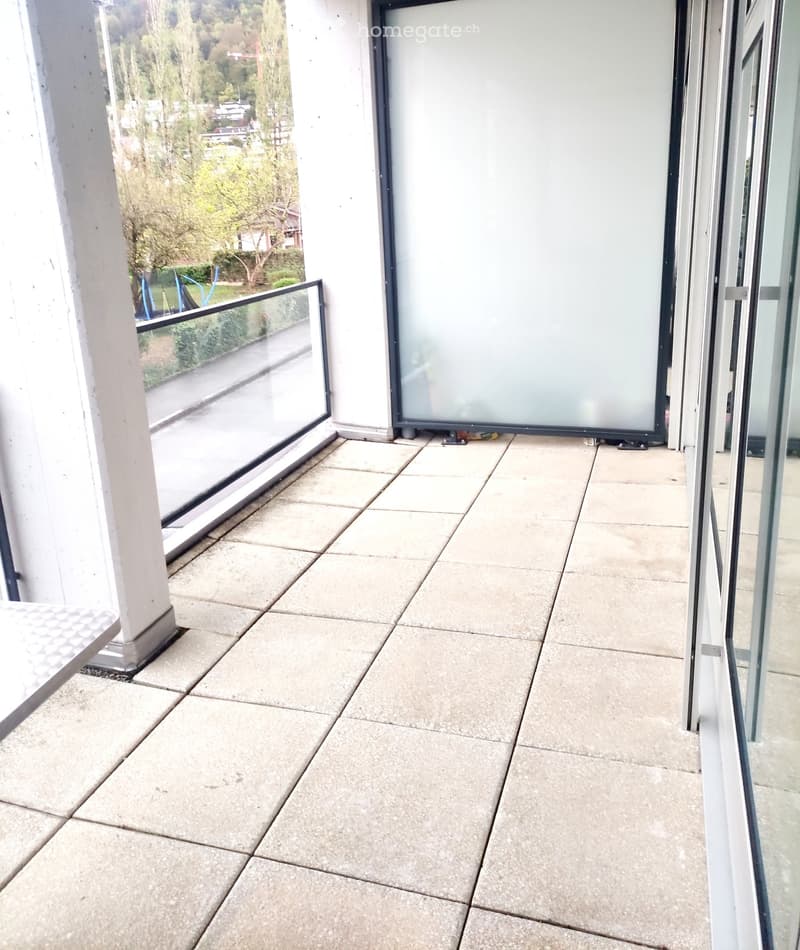 Balcony with tiled floor, glass railings, and a view of a landscaped area with trees and a road.