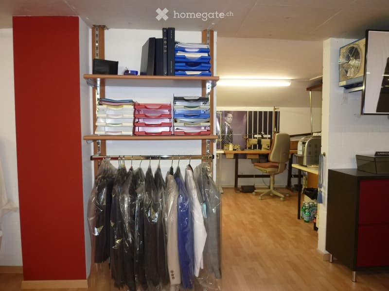 Office space with wooden floor, shelves filled with books and files, clothing rack, desk with chair, and a fan on the wall.