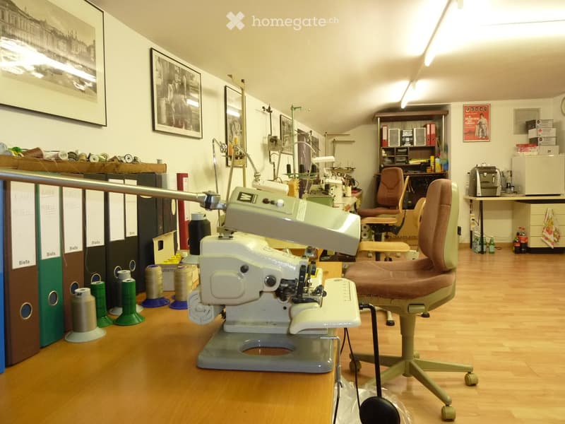 A well-lit room with wooden flooring, featuring a sewing machine, chairs, a table, filing cabinets, and shelves with books and supplies.