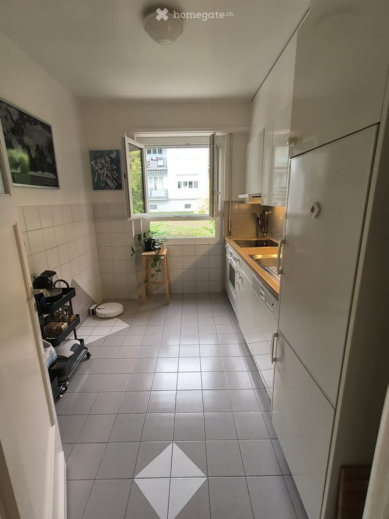 Modern kitchen with white cabinets, stainless steel appliances, tiled floor, and a small window with a view of the building outside. Includes a small stool with a plant, a cart with various items, and framed pictures on the wall.