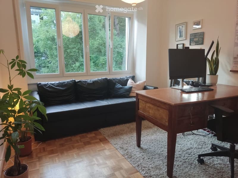 Bright living room with large windows, a black leather sofa, a wooden desk with a computer setup, and potted plants.