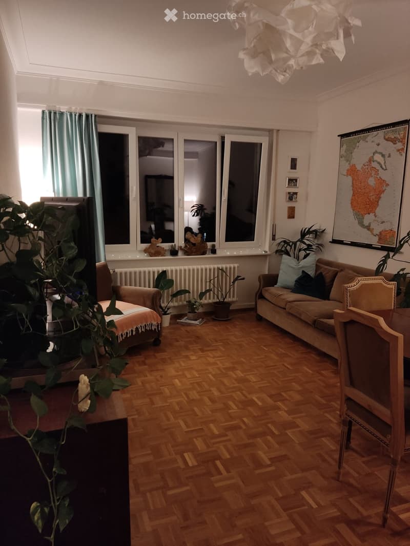 Living room with wooden parquet flooring, large windows with blue curtains, a beige sofa with cushions, a wooden armchair, a small table with a plant, and a large map on the wall.
