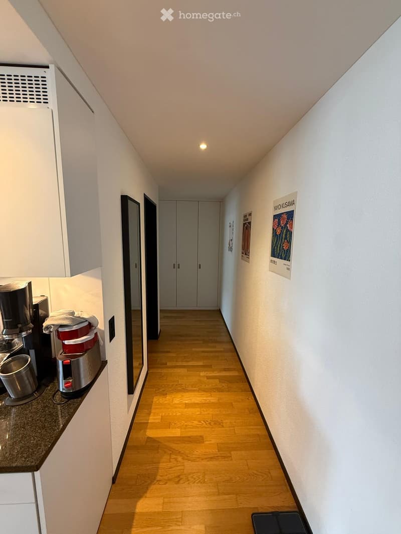 A narrow hallway with wooden flooring, white walls, and several doors leading to different rooms. There is a small counter area with a coffee maker and other appliances on the left side.