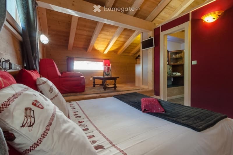 Cozy wooden-beamed bedroom with a large bed, red armchair, small table, and television mounted on the wall. The room features exposed wooden beams and warm lighting.