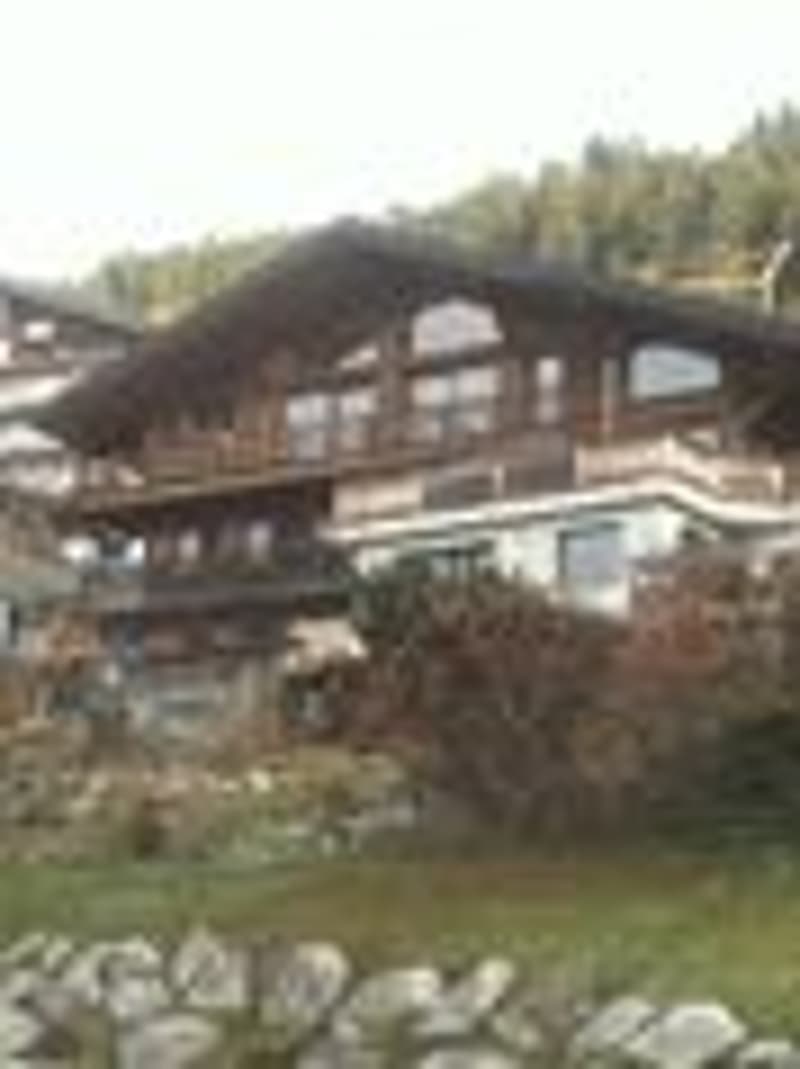 A large, multi-story wooden chalet-style building with a steeply pitched roof, surrounded by trees and a stone pathway.