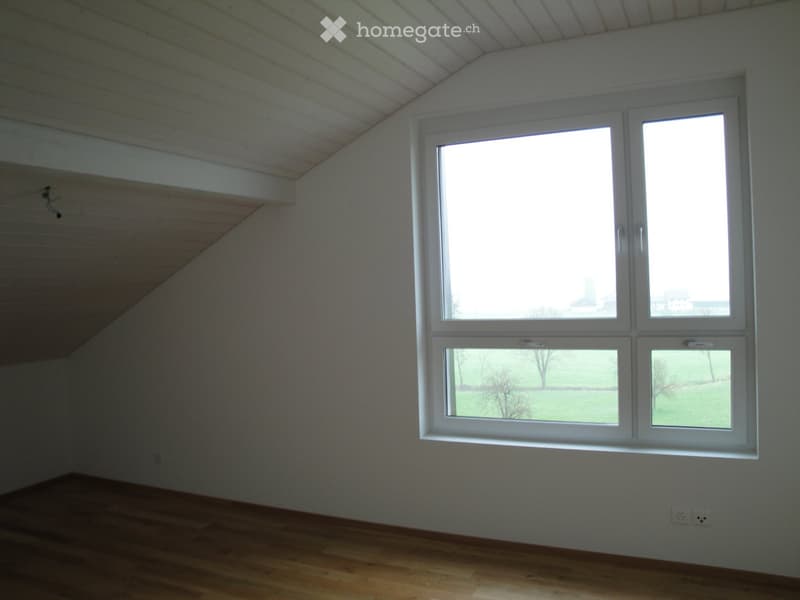 Empty room with wooden flooring, white walls, and large windows offering a view of a green landscape.