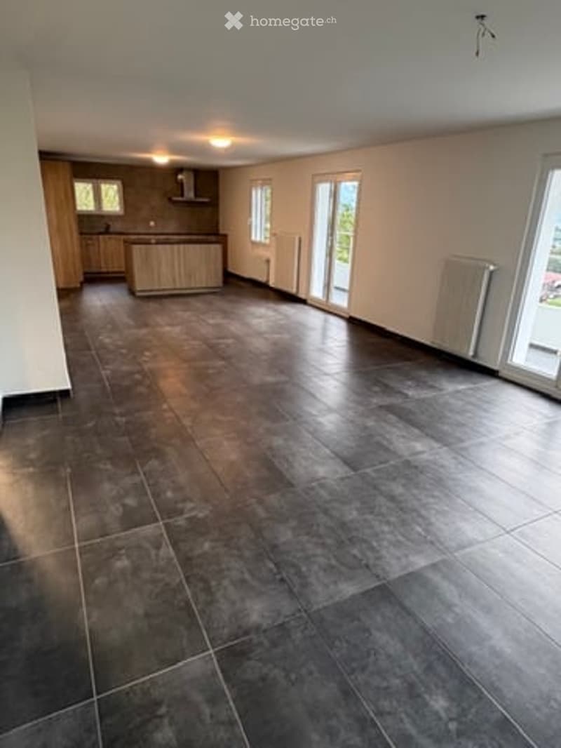 Empty room with dark grey tiled floor, white walls, and large windows with sliding doors leading to an outdoor area. The room is unfurnished, showing only the basic structure and flooring.