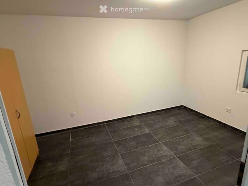 Empty room with light-colored walls, dark tiled floor, and a small window on one wall.
